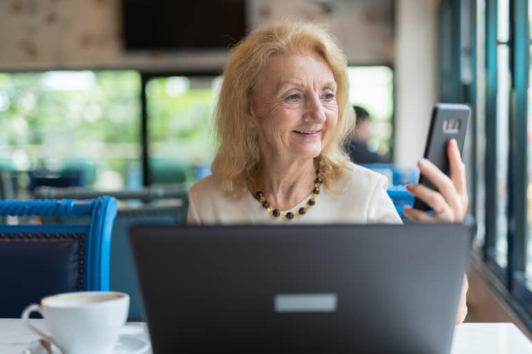 Happy senior woman smiling and while using phone and laptop computer in restaurant